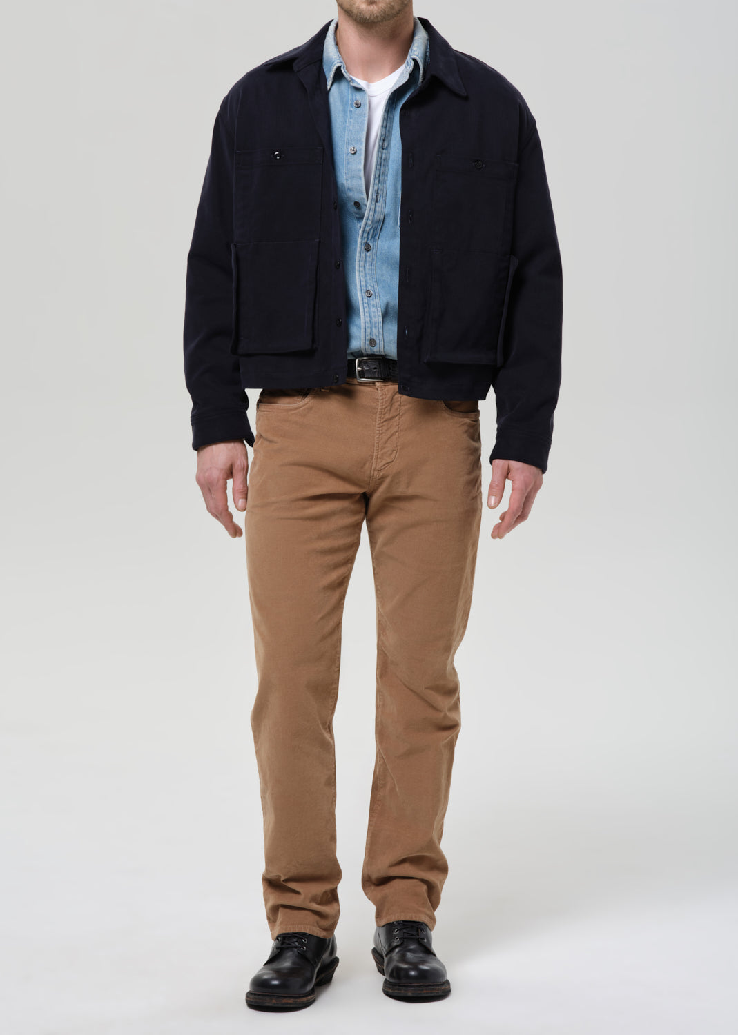 Person wearing a black jacket, light blue shirt, and brown pants on a grey background