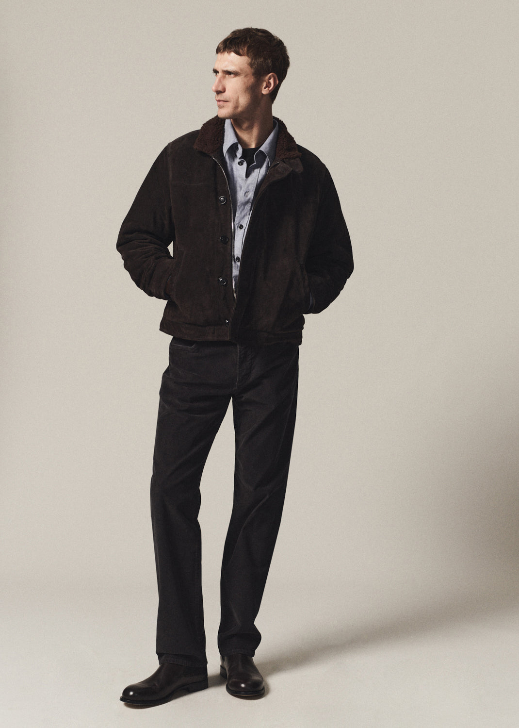 A man wearing a dark brown suede jacket over a light blue button-down shirt, paired with black trousers and black leather shoes, standing against a neutral beige studio background.