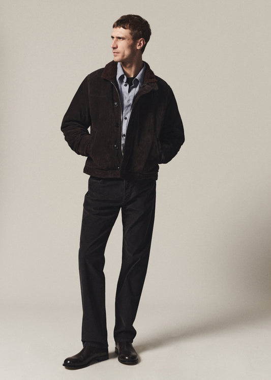 A man wearing a dark brown suede jacket over a light blue button-down shirt, paired with black trousers and black leather shoes, standing against a neutral beige studio background.