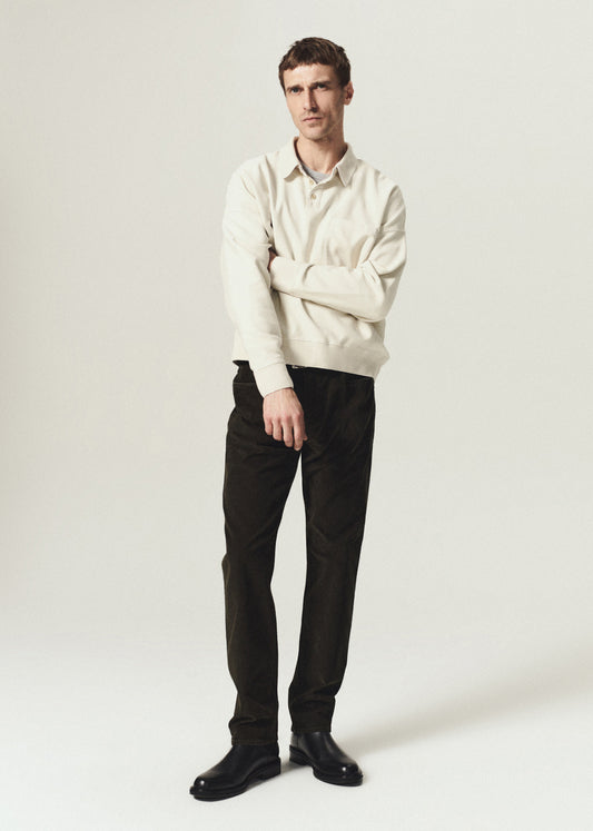 A man modeling a cream-colored long-sleeve knit polo sweater over a collared shirt, paired with olive trousers and black leather shoes, against a plain light gray studio background.