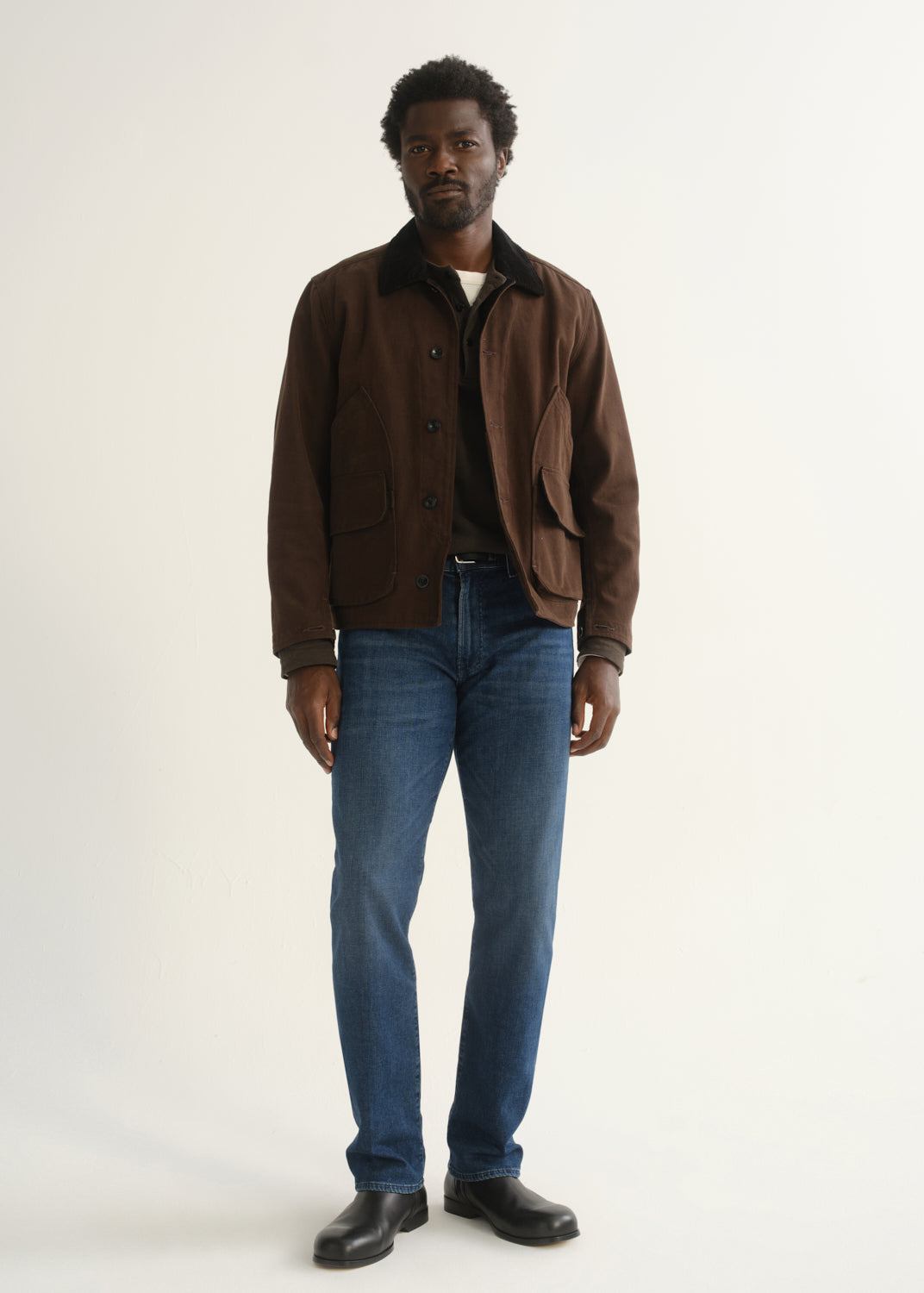 Person wearing medium blue straight leg jeans with a brown jacket, dark shirt, and black shoes, front full body view on a light grey background.
