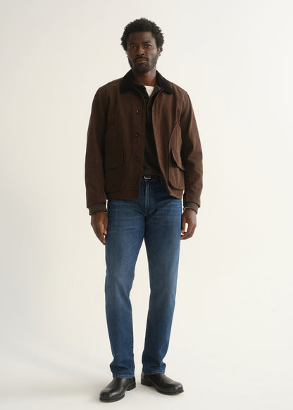 Person wearing medium blue straight leg jeans with a brown jacket, dark shirt, and black shoes, front full body view on a light grey background.
