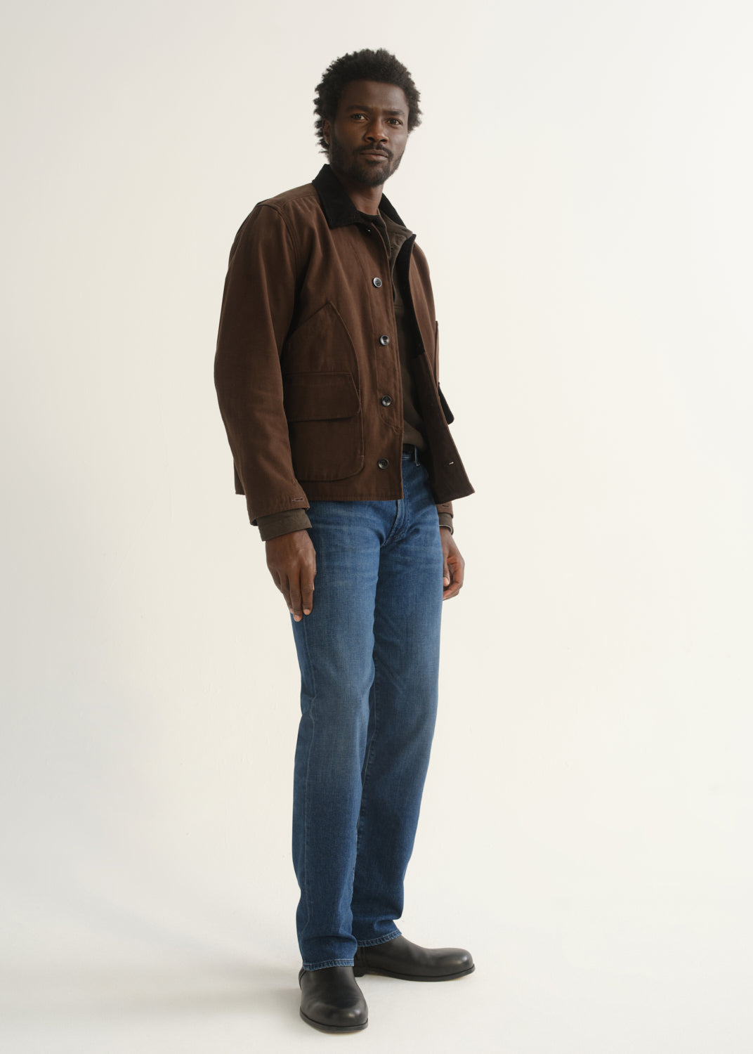 Person wearing medium blue straight leg jeans with a brown jacket, dark shirt, and black shoes, angled front full body view on a light grey background.