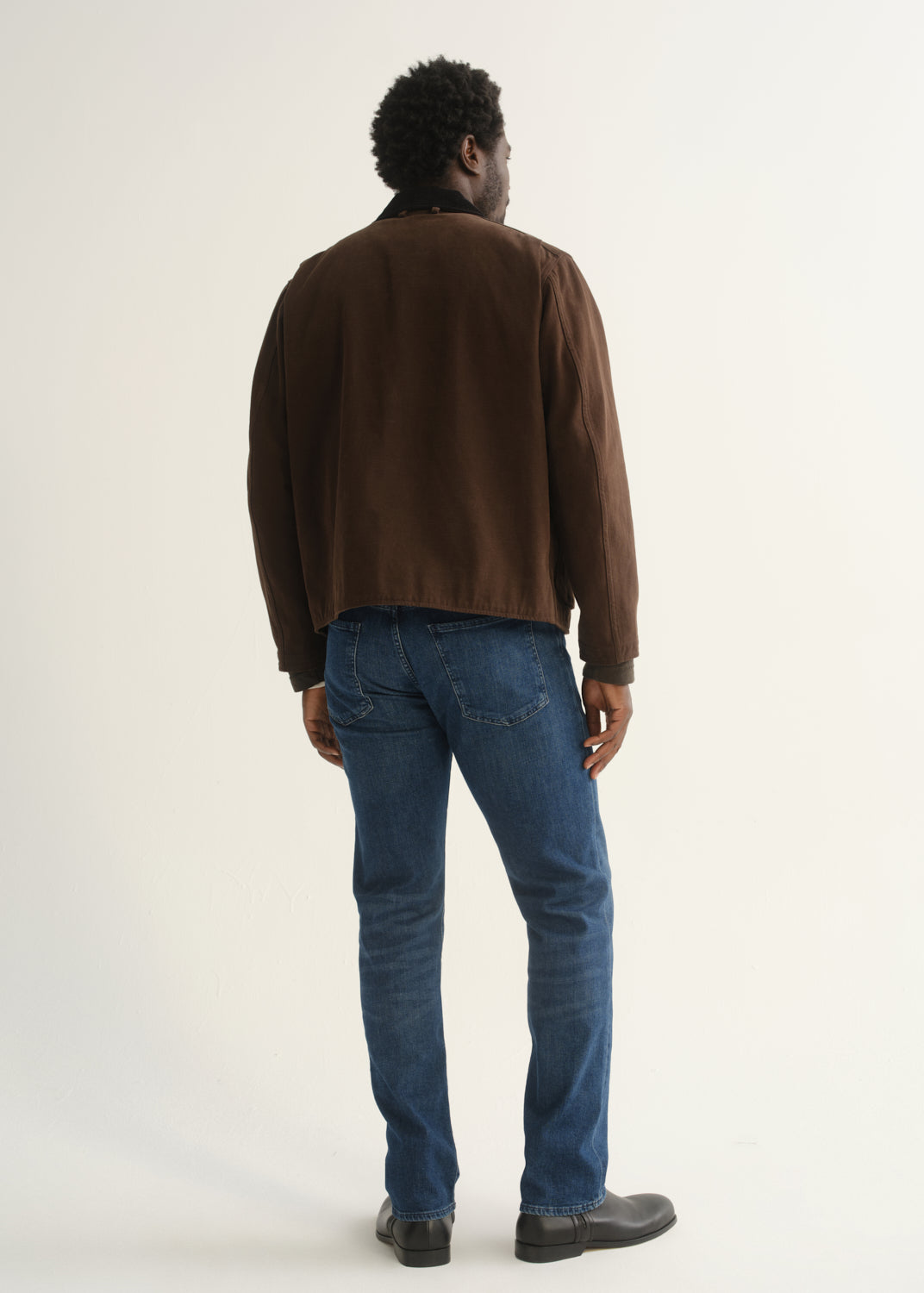 Person wearing medium blue straight leg jeans with a brown jacket and black shoes, back full body view on a light grey background.