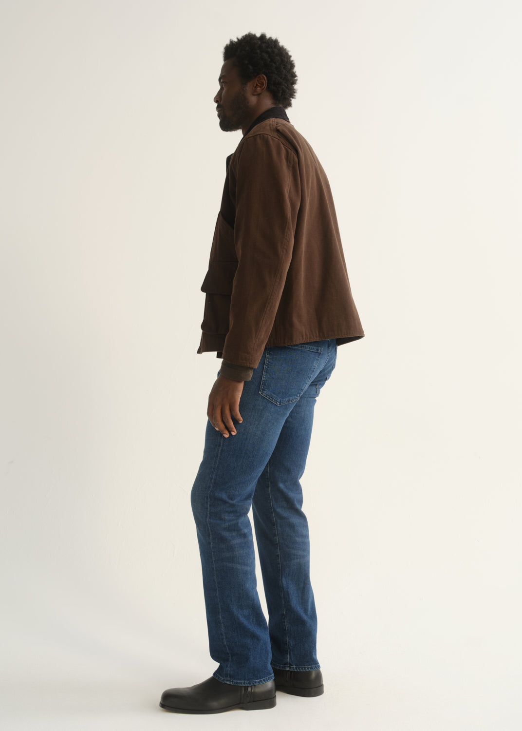 Person wearing medium blue straight leg jeans with a brown jacket and black shoes, side full body view on a light grey background.