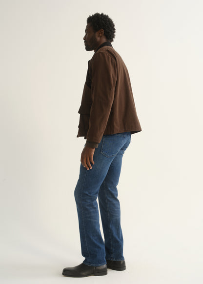 Person wearing medium blue straight leg jeans with a brown jacket and black shoes, side full body view on a light grey background.