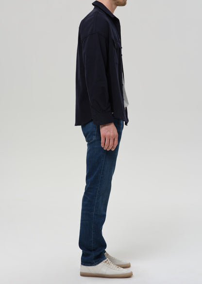Person wearing medium blue straight leg jeans with a navy bomber jacket and light grey crewneck tee viewed from the side on a grey background.