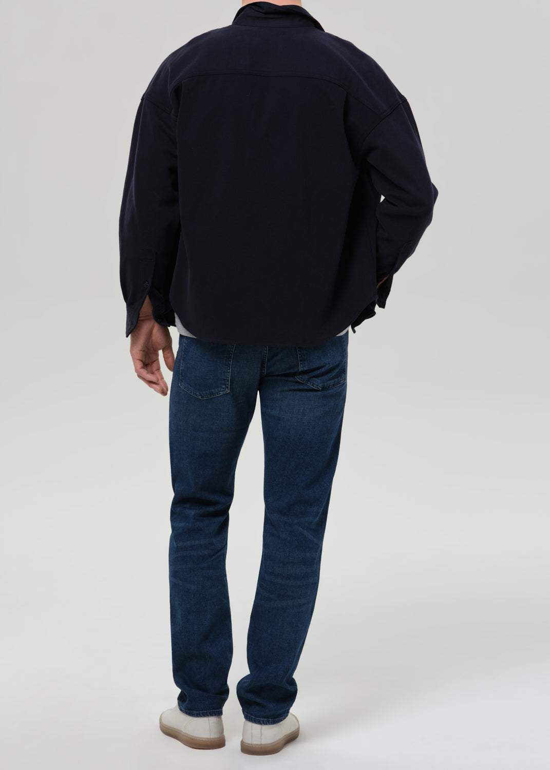Person wearing medium blue straight leg jeans with a navy bomber jacket and light grey crewneck tee viewed from the back on a grey background.