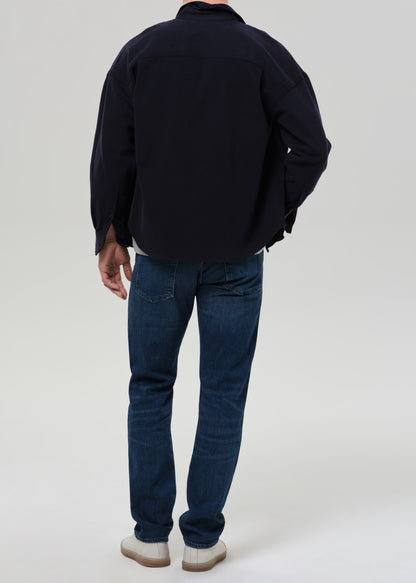 Person wearing medium blue straight leg jeans with a navy bomber jacket and light grey crewneck tee viewed from the back on a grey background.