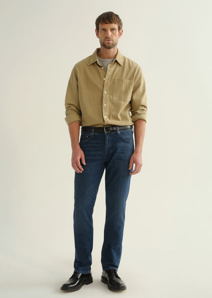 Person wearing dark blue straight leg jeans with a khaki long sleeve button-up shirt and black boots, front full body view on a light grey background.