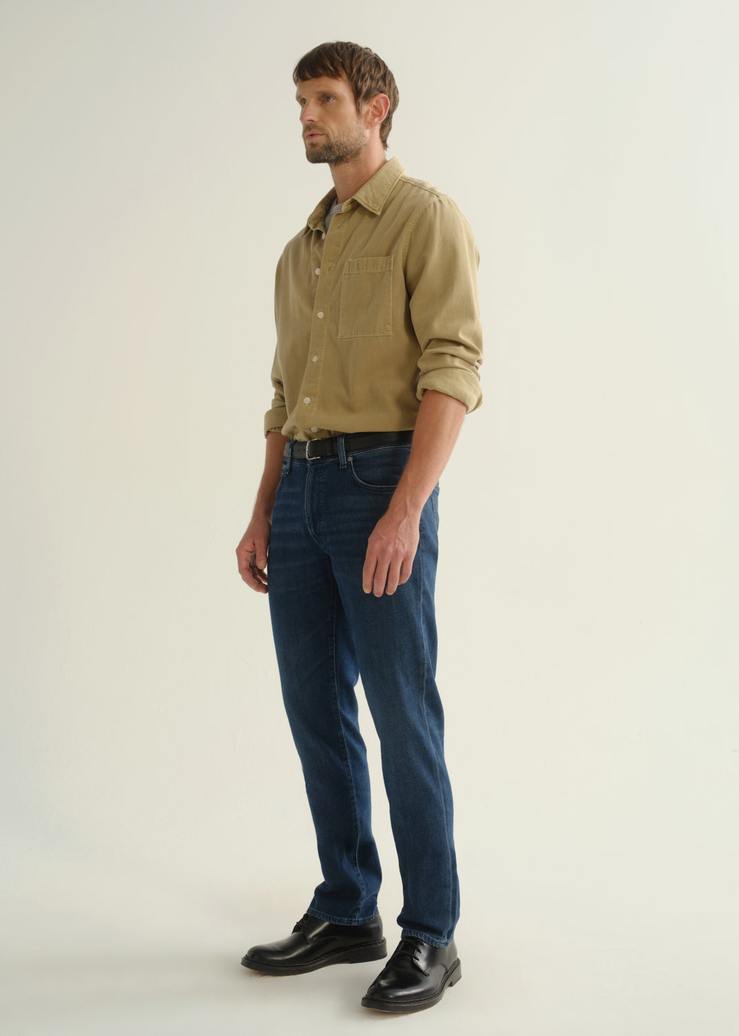 Person wearing dark blue straight leg jeans with a khaki long sleeve button-up shirt and black boots, angled front full body view on a light grey background.
