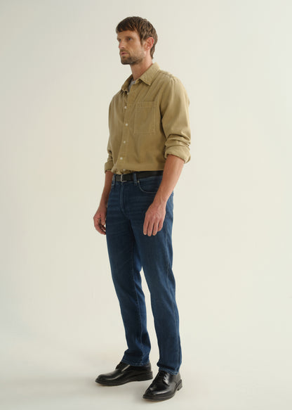Person wearing dark blue straight leg jeans with a khaki long sleeve button-up shirt and black boots, angled front full body view on a light grey background.