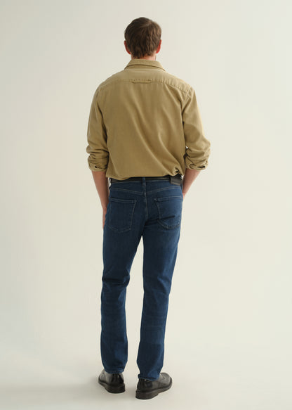 Person wearing dark blue straight leg jeans with a khaki long sleeve button-up shirt and black boots, back full body view on a light grey background.