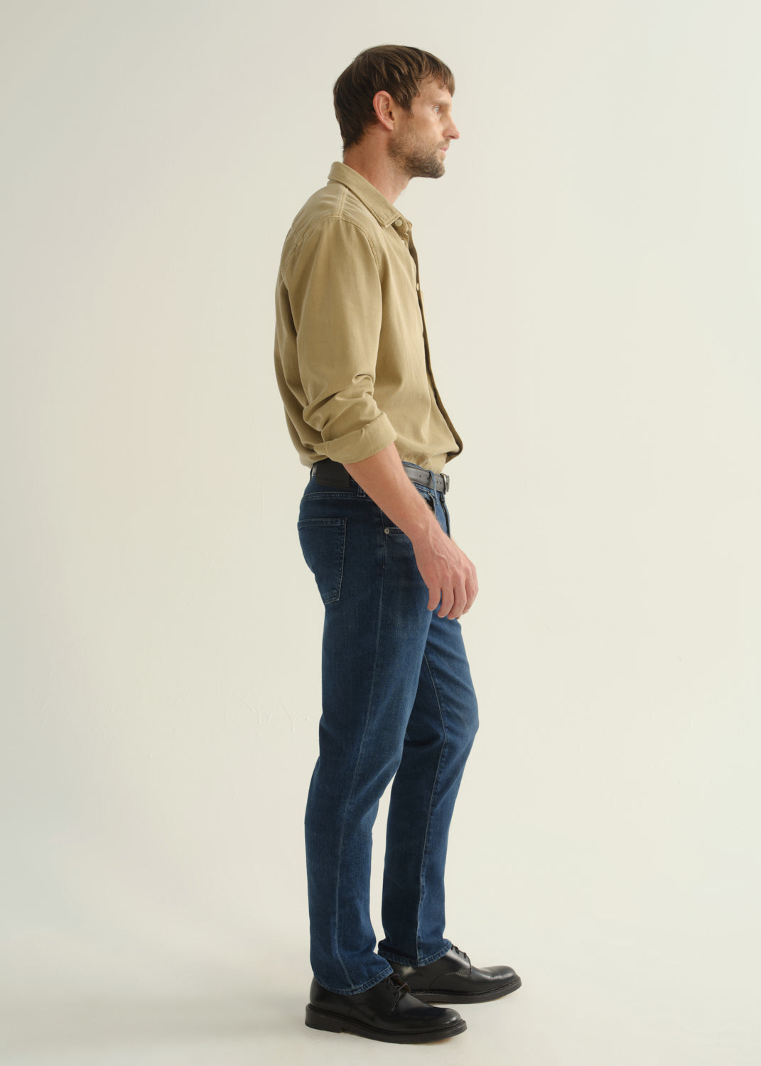 Person wearing dark blue straight leg jeans with a khaki long sleeve button-up shirt and black boots, side full body view on a light grey background.