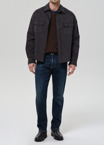 Person wearing dark indigo straight leg jeans with a charcoal chore jacket and dark brown crewneck tee on a grey background.