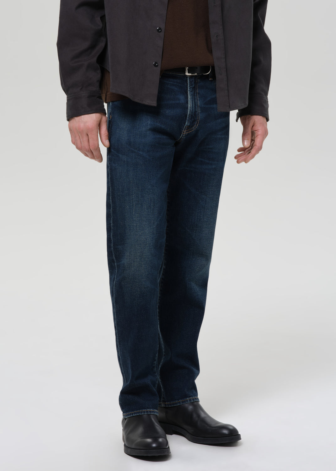 Person wearing dark indigo straight leg jeans with a charcoal chore jacket and dark brown crewneck tee on a grey background.