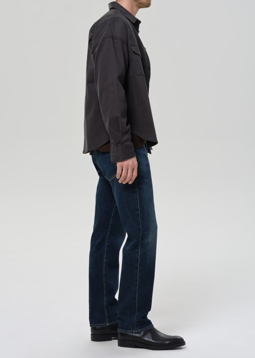 Person wearing dark indigo straight leg jeans with a charcoal chore jacket and dark brown crewneck tee viewed from the side on a grey background.