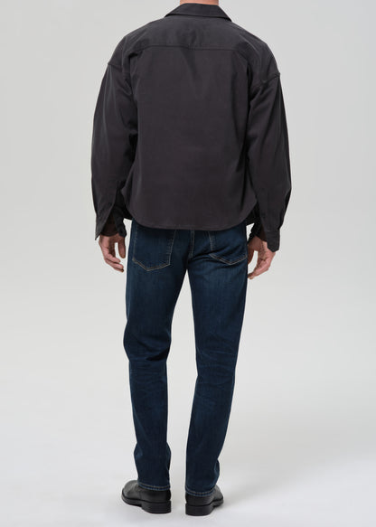 Person wearing dark indigo straight leg jeans with a charcoal chore jacket and dark brown crewneck tee viewed from the back on a grey background.