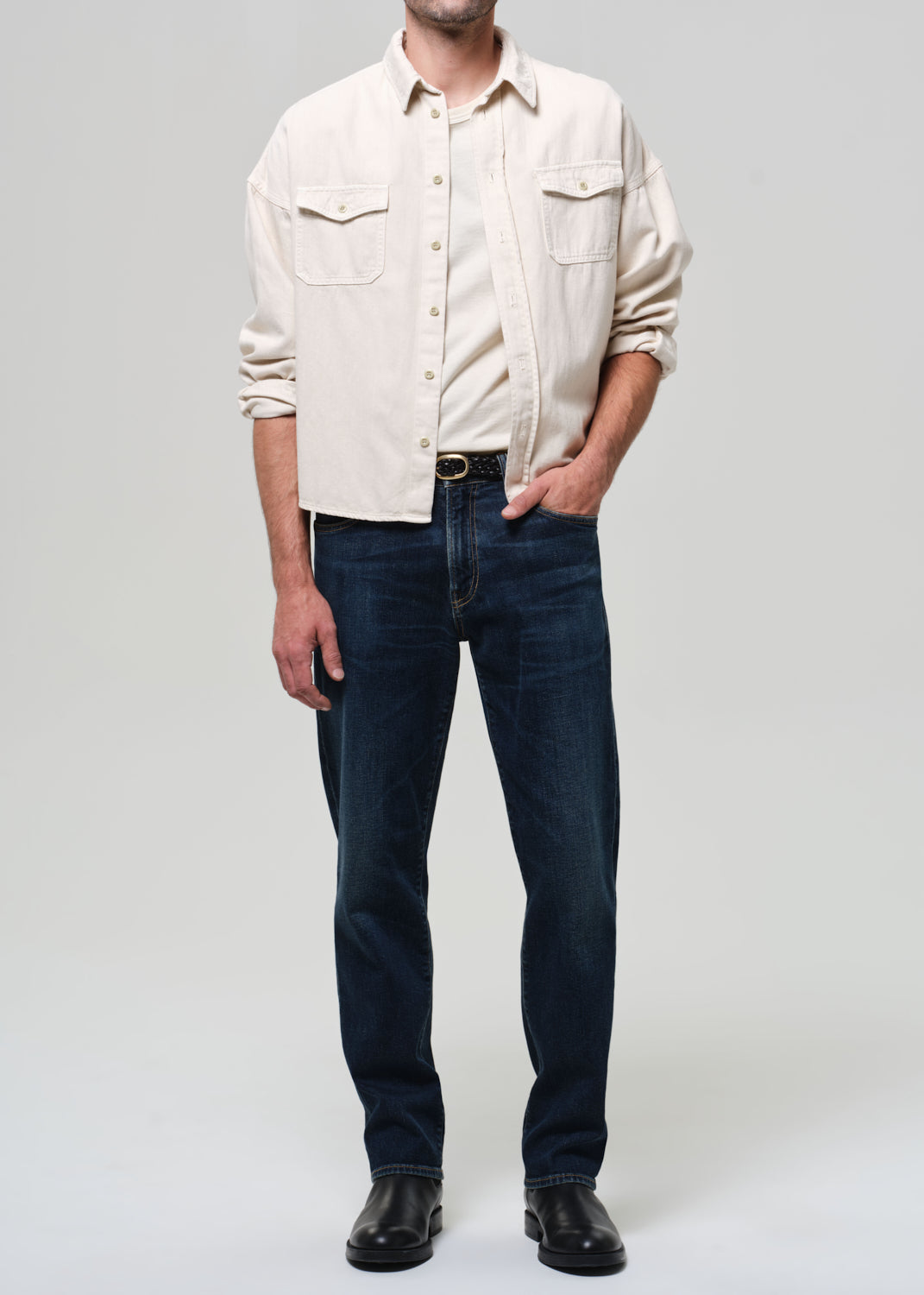 Man wearing a beige shirt and dark jeans on a plain background