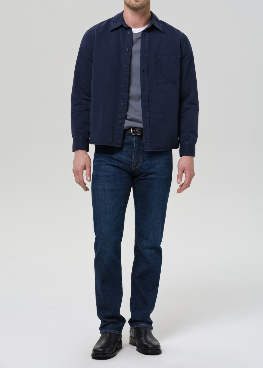 Man wearing a navy shirt, gray tee, and dark jeans on a plain background