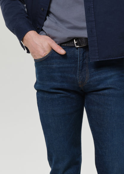 Man wearing a navy shirt, gray tee, and dark jeans on a plain background