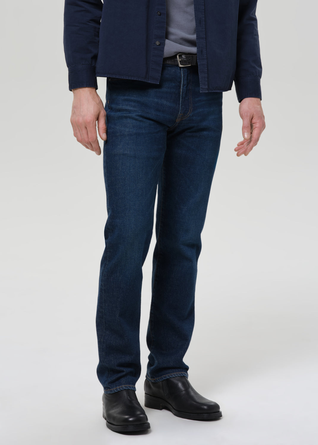 Man wearing a navy shirt, gray tee, and dark jeans on a plain background
