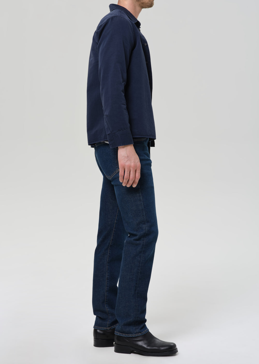 Man wearing a navy shirt, gray tee, and dark jeans on a plain background