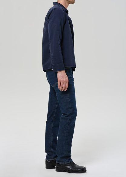 Man wearing a navy shirt, gray tee, and dark jeans on a plain background