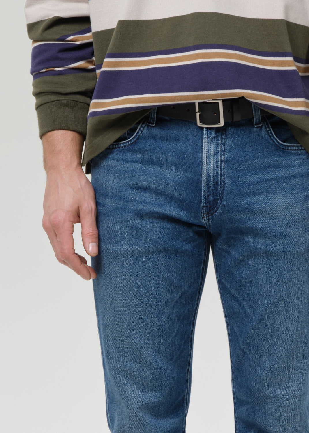 A close-up of a person wearing blue jeans and a green, navy, and beige striped long-sleeve shirt, cropped from the chest down against a grey background.