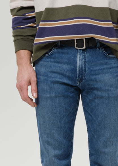 A close-up of a person wearing blue jeans and a green, navy, and beige striped long-sleeve shirt, cropped from the chest down against a grey background.