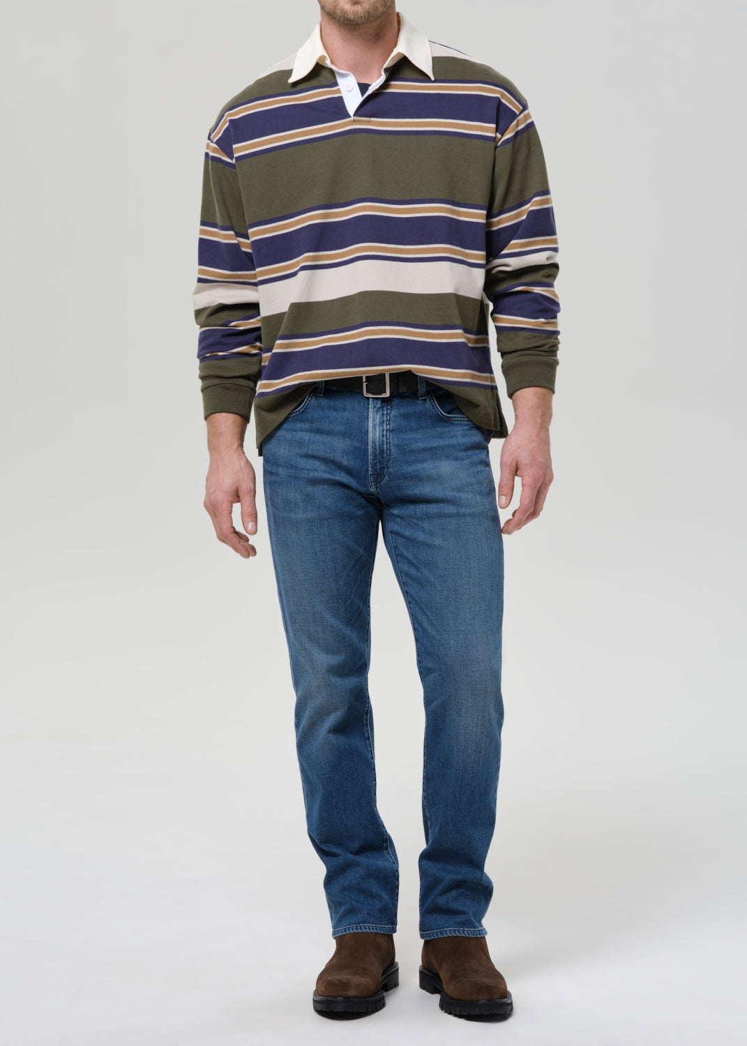 A man wearing a green, navy, and beige striped long-sleeve rugby shirt, blue jeans, and brown suede boots, standing against a grey background.