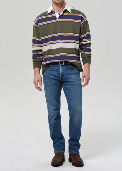A man wearing a green, navy, and beige striped long-sleeve rugby shirt, blue jeans, and brown suede boots, standing against a grey background.