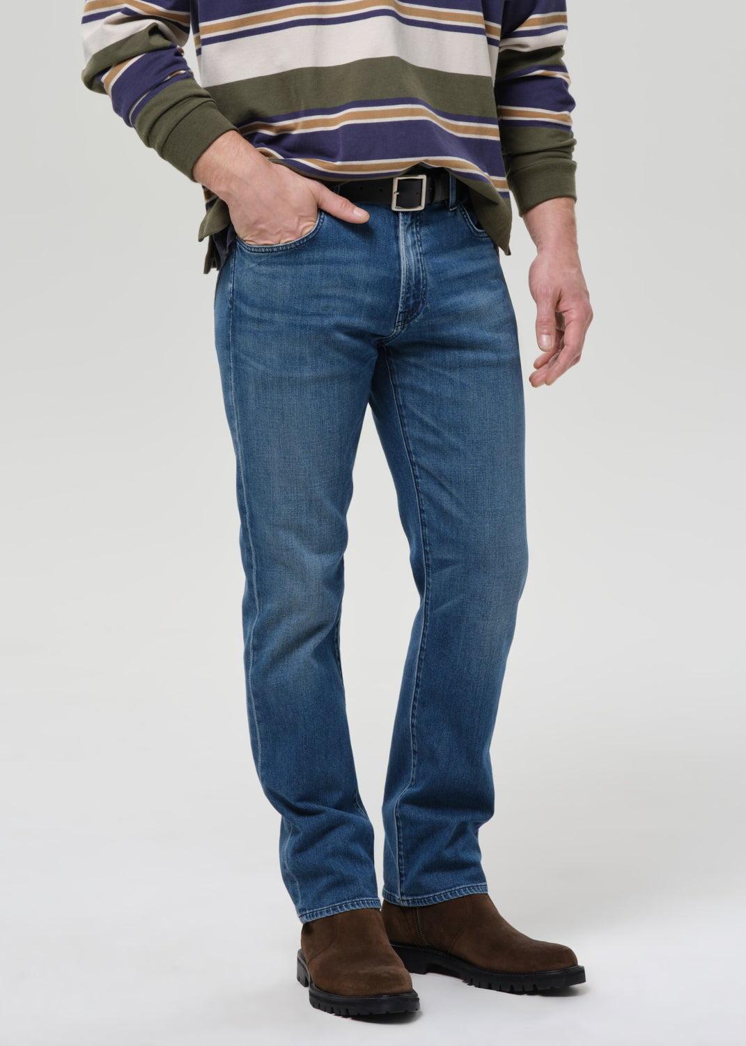 A man wearing blue straight-leg jeans, a green and navy striped sweater, and brown suede boots, standing against a grey background.