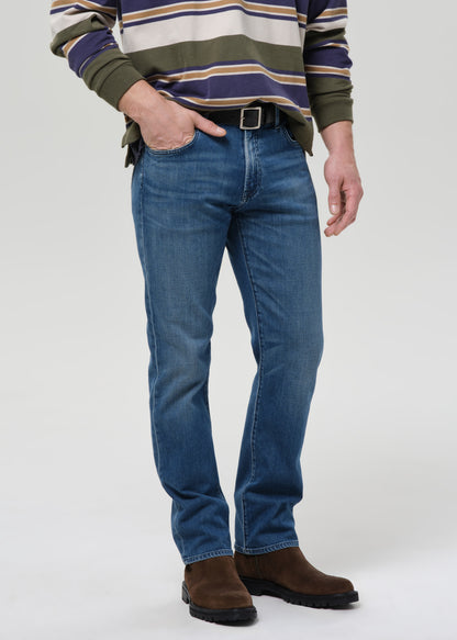 A man wearing blue straight-leg jeans, a green and navy striped sweater, and brown suede boots, standing against a grey background.