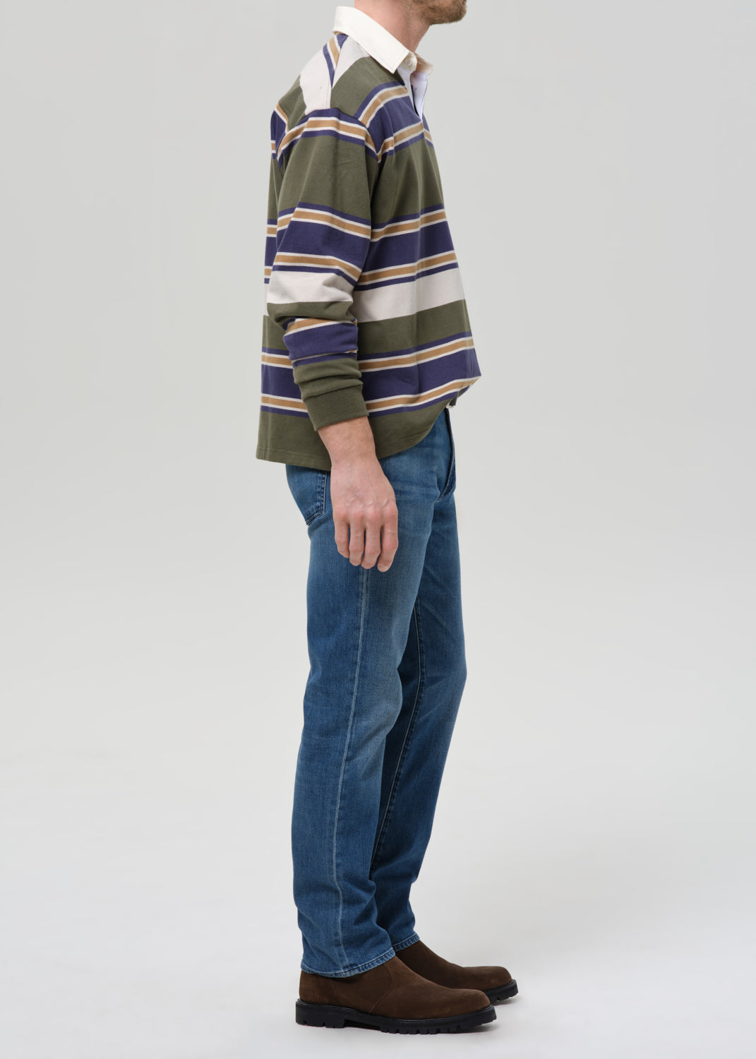 A man shown in profile wearing a green, navy, and beige striped long-sleeve rugby shirt, blue jeans, and brown suede boots against a grey background.