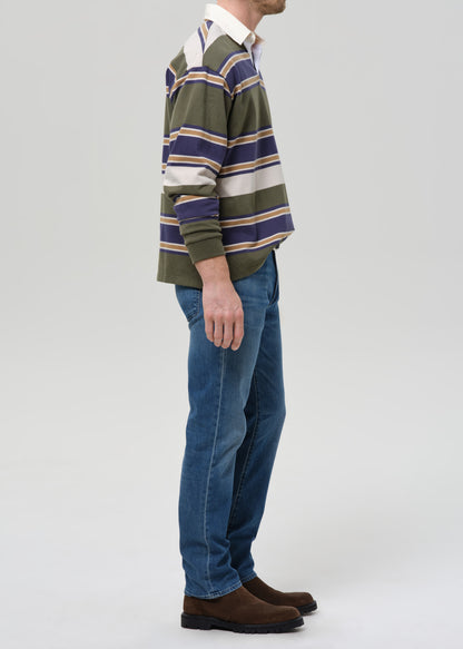A man shown in profile wearing a green, navy, and beige striped long-sleeve rugby shirt, blue jeans, and brown suede boots against a grey background.