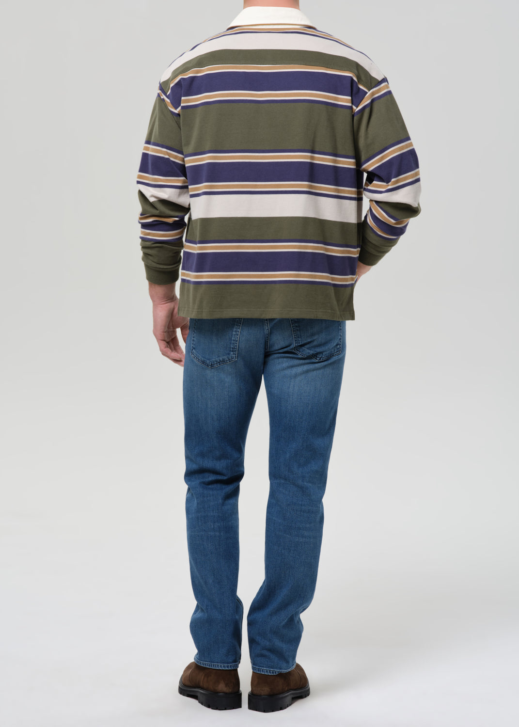 A man viewed from the back wearing a long-sleeve striped rugby shirt in olive green, navy blue, and cream, paired with blue jeans and brown shoes, against a grey background.