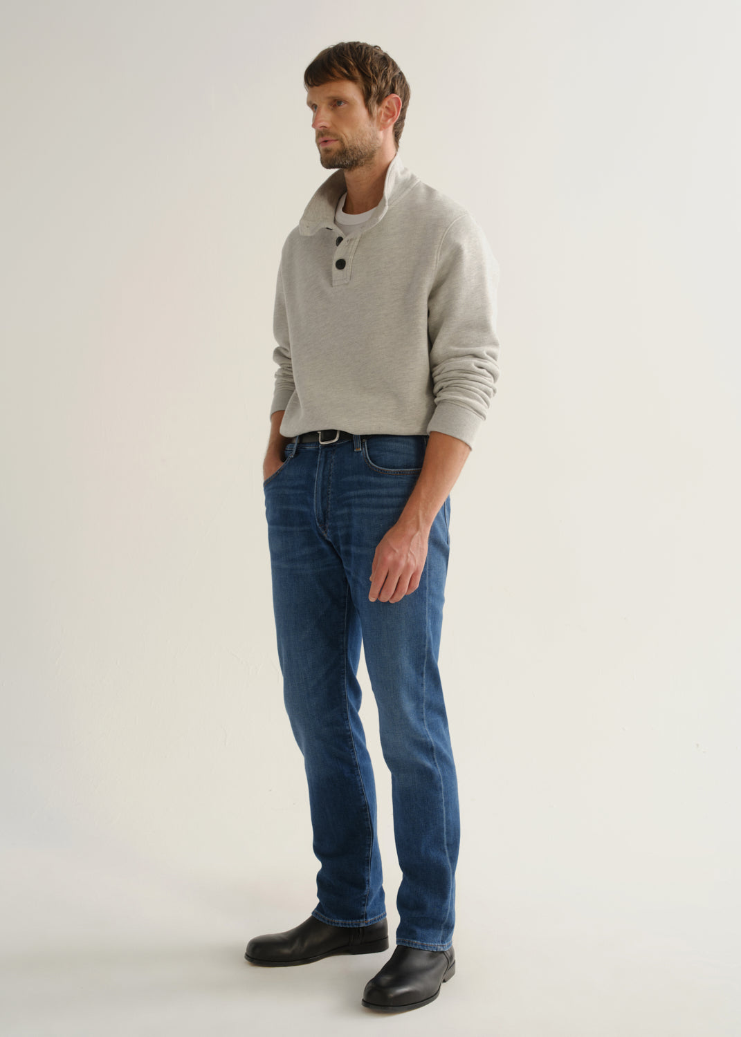 Person wearing medium blue straight leg jeans with a light grey long sleeve collared pullover and black boots, angled front full body view on a light grey background.