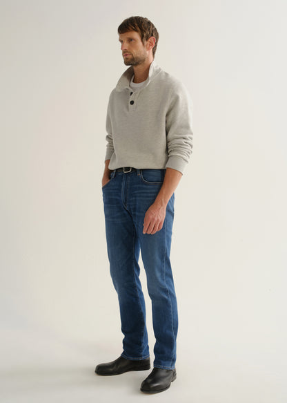 Person wearing medium blue straight leg jeans with a light grey long sleeve collared pullover and black boots, angled front full body view on a light grey background.