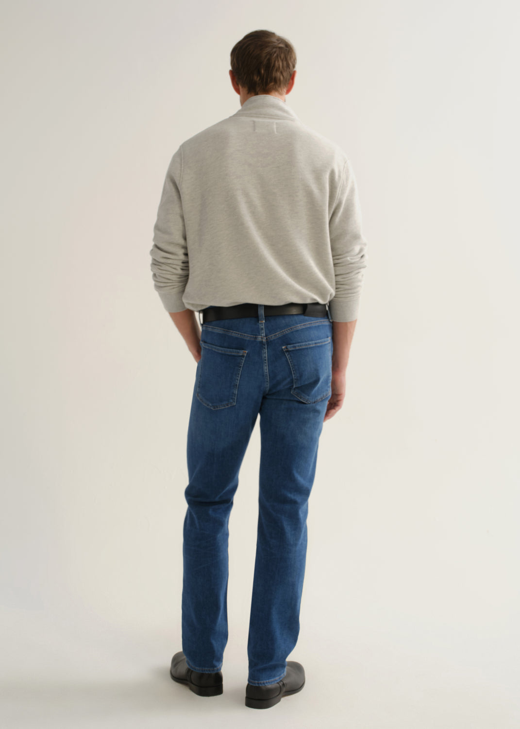 Person wearing medium blue straight leg jeans with a light grey long sleeve collared pullover and black boots, back full body view on a light grey background.