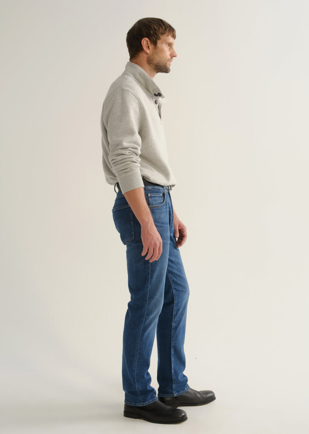 Person wearing medium blue straight leg jeans with a light grey long sleeve collared pullover and black boots, side full body view on a light grey background.