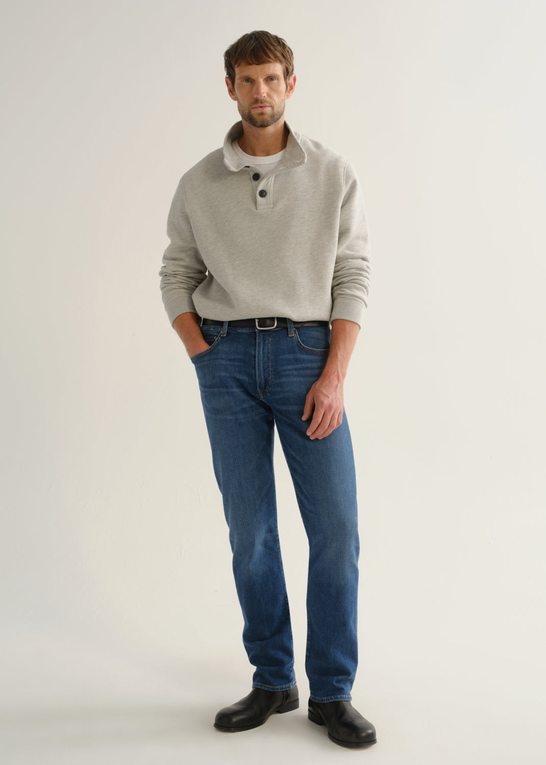 Person wearing medium blue straight leg jeans with a light grey long sleeve collared pullover and black boots, front full body view on a light grey background.