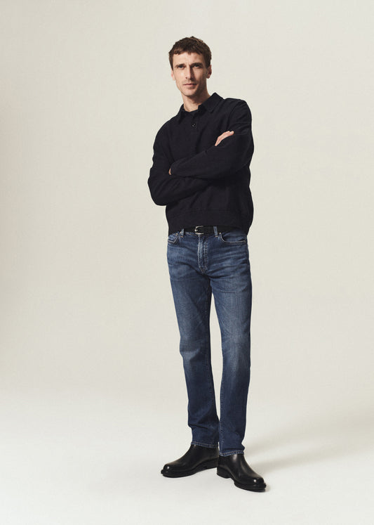 A man stands with arms crossed wearing a black long-sleeve polo shirt, blue jeans, and black leather boots against a plain light beige studio background.