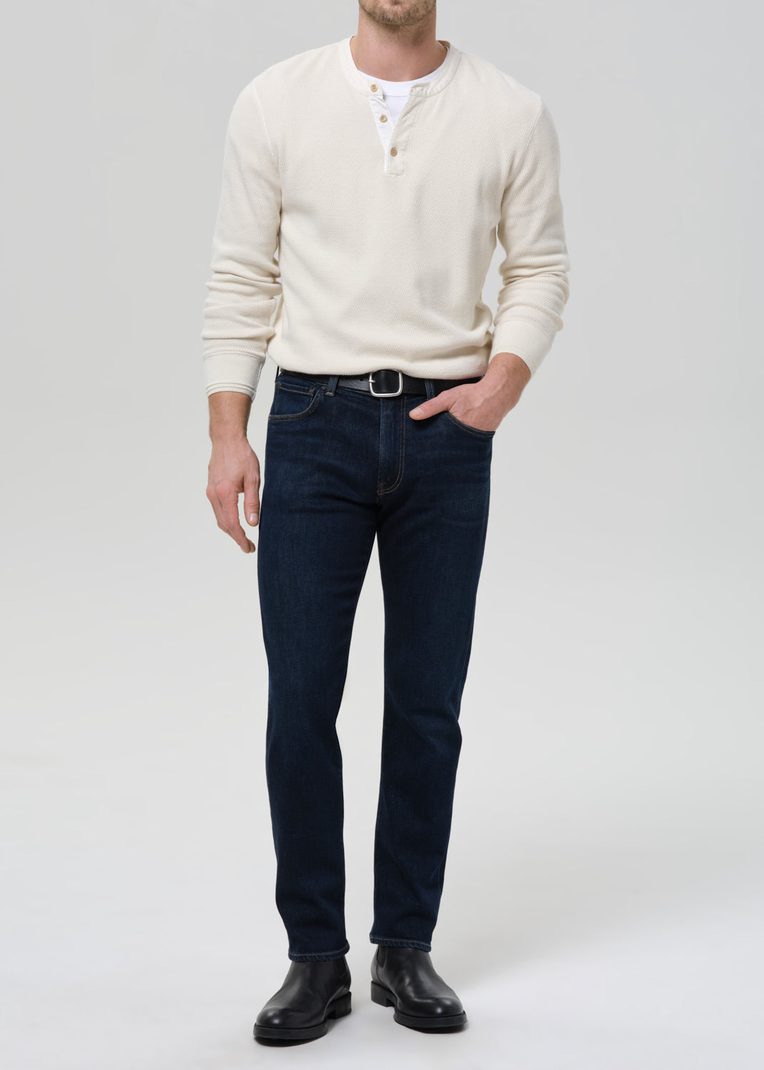 A man wearing a cream-colored long-sleeve henley shirt, dark blue slim-fit jeans, a black belt, and black leather boots, standing against a grey background.