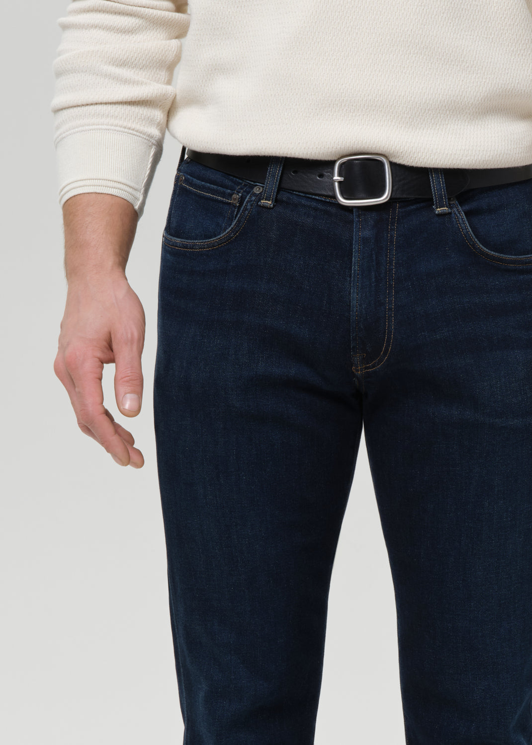 A close-up of a person wearing dark blue denim jeans with a black leather belt and silver buckle, paired with a cream ribbed sweater, against a grey background.