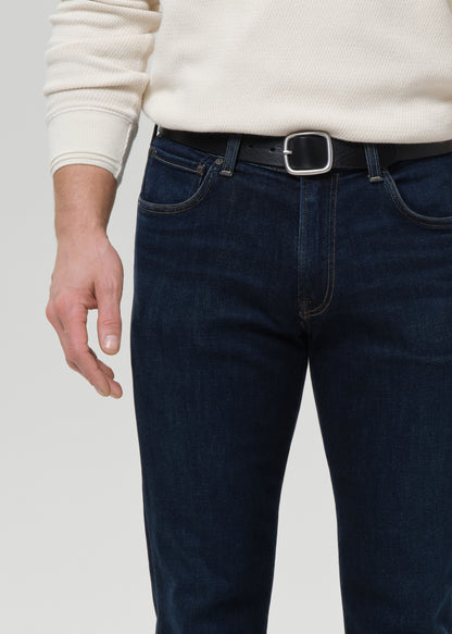 A close-up of a person wearing dark blue denim jeans with a black leather belt and silver buckle, paired with a cream ribbed sweater, against a grey background.