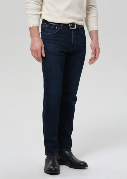 A person wearing dark blue slim-fit jeans with a black belt and black leather boots, standing against a grey background.