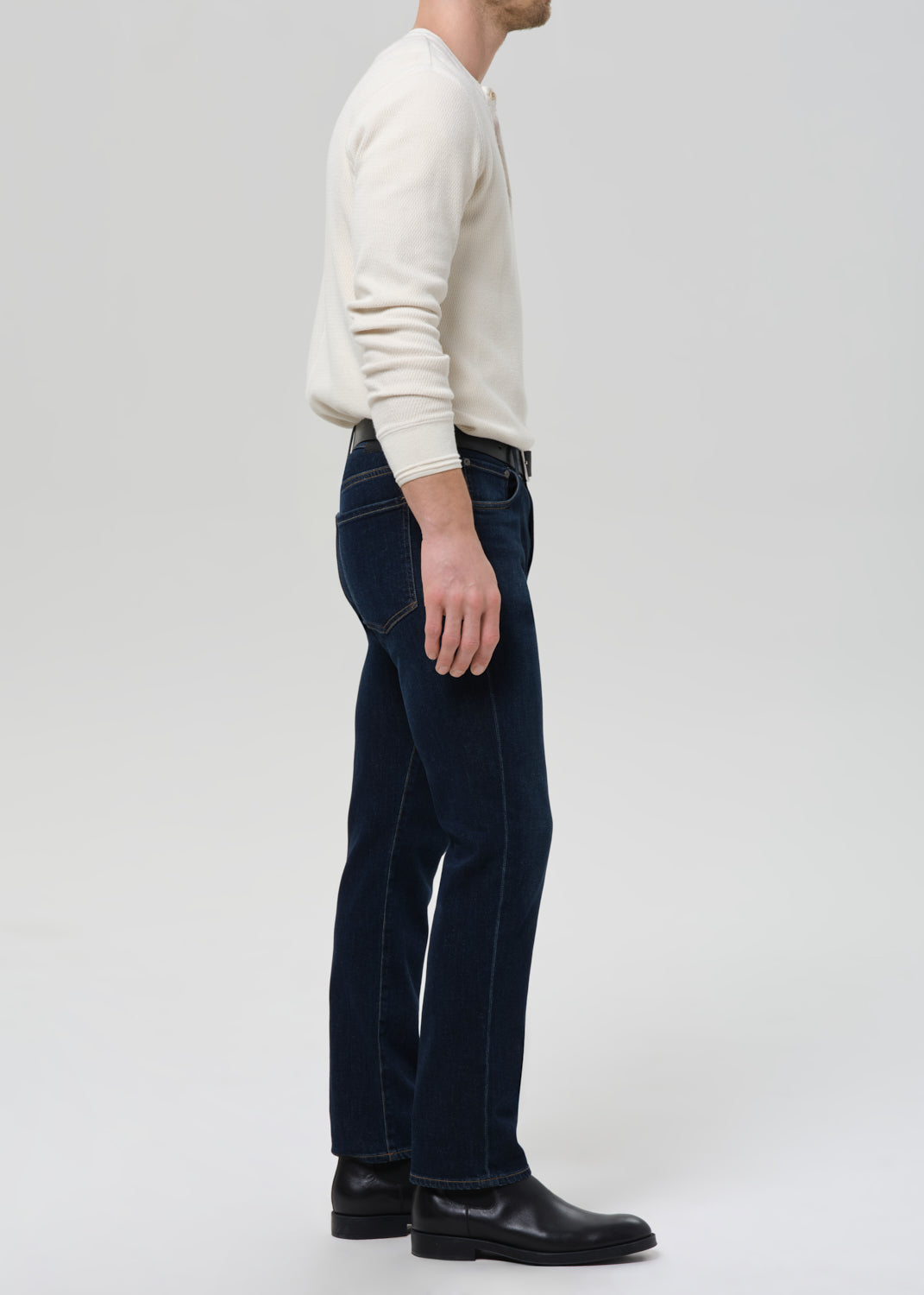 A man shown in profile wearing dark blue straight-leg jeans, a cream long-sleeve knit sweater, and black leather boots against a grey background.