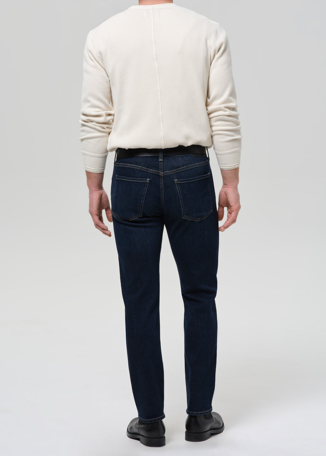 A man viewed from behind wearing dark navy blue slim-fit jeans, a cream-colored long-sleeve knit sweater, and black leather shoes, set against a grey background.