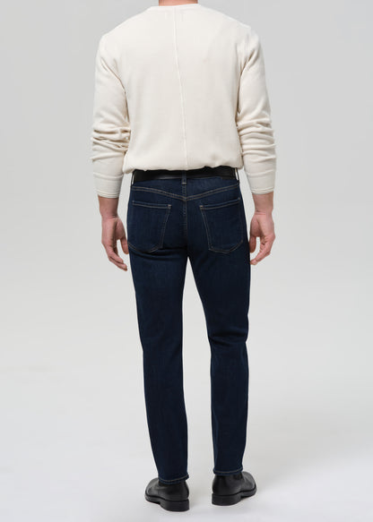 A man viewed from behind wearing dark navy blue slim-fit jeans, a cream-colored long-sleeve knit sweater, and black leather shoes, set against a grey background.